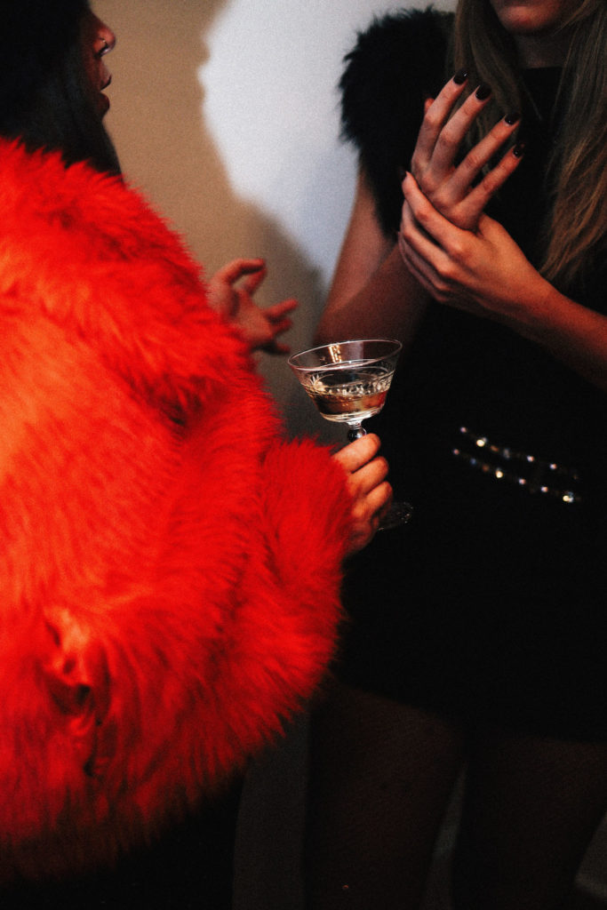 Two women in stylish dresses have a conversation, one holding a cocktail glass, creating a chic party atmosphere.