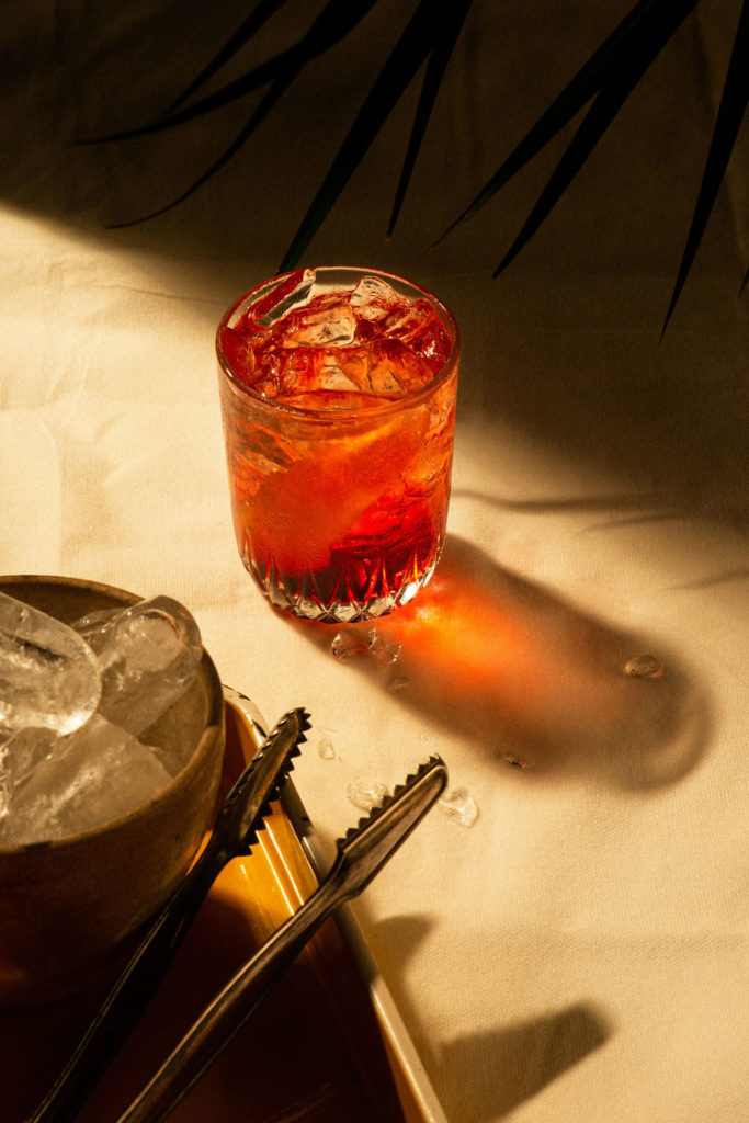 Chilled cocktail with orange slice in sunlight, next to ice bowl and tongs on a table.